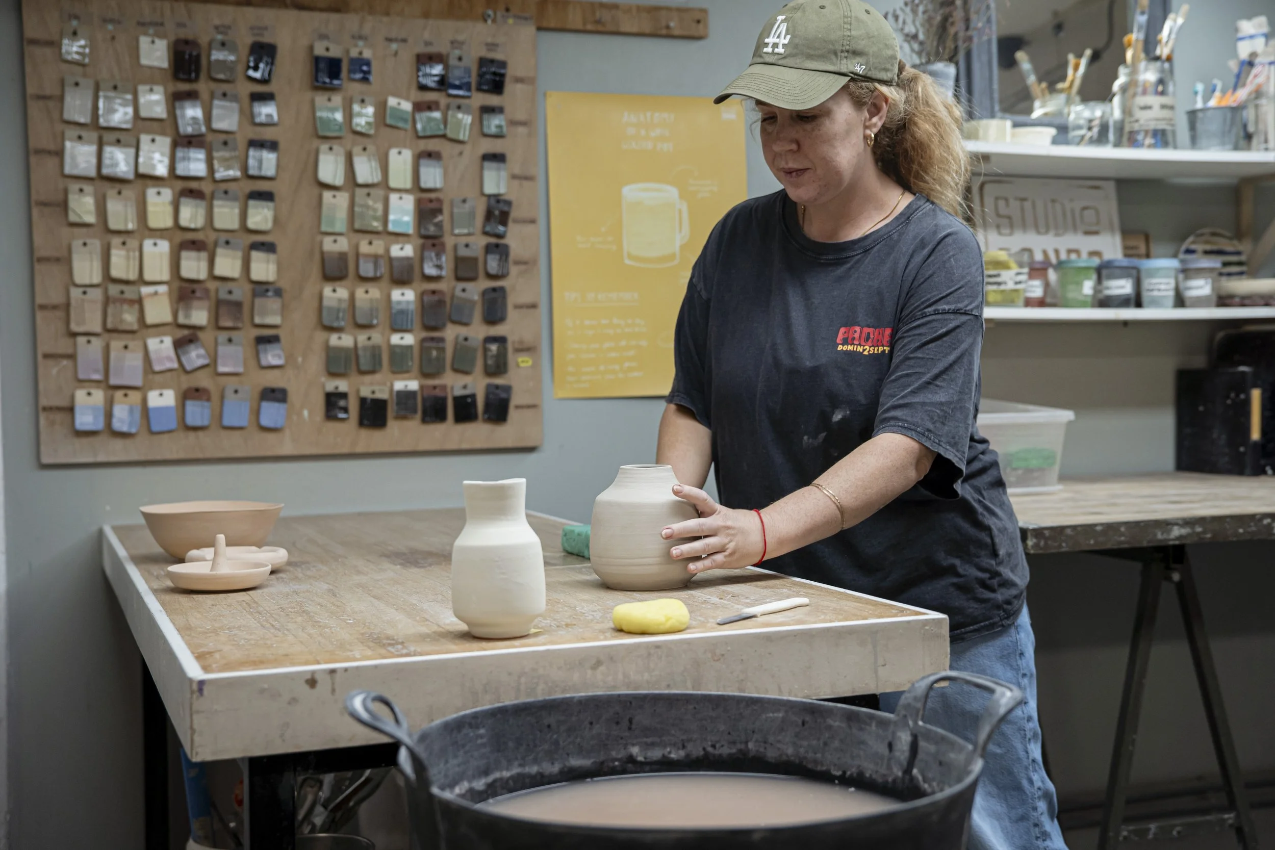 A woman with curly hair wearing a black T-shirt and a baseball cap is shaping a ceramic vase in a pottery studio.