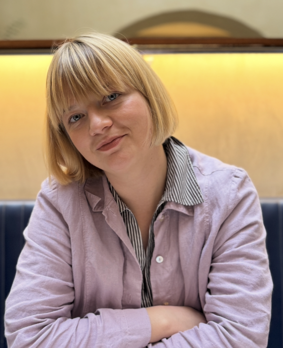 A woman with blonde bob hair and blue eyes sitting at a table, with folded arms, wearing a lavender jacket over a striped shirt, inside a restaurant or cafe.