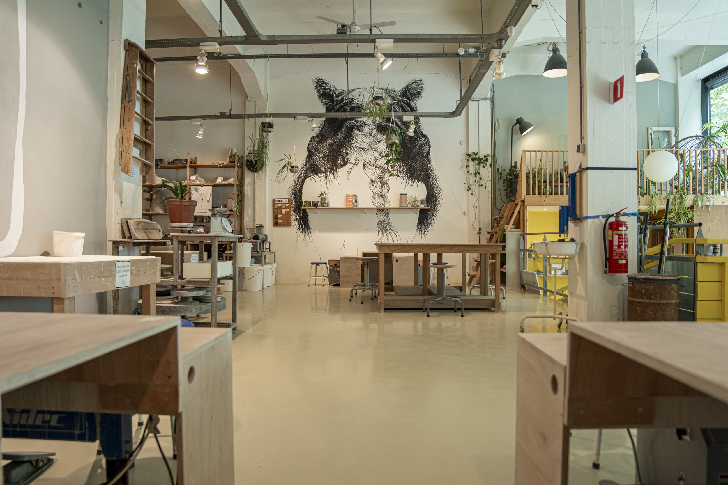 Interior of a modern cafe or workspace with a large wall mural of a gorilla painted in black and white. Wooden furniture, plants, and lighting fixtures are visible throughout the space.