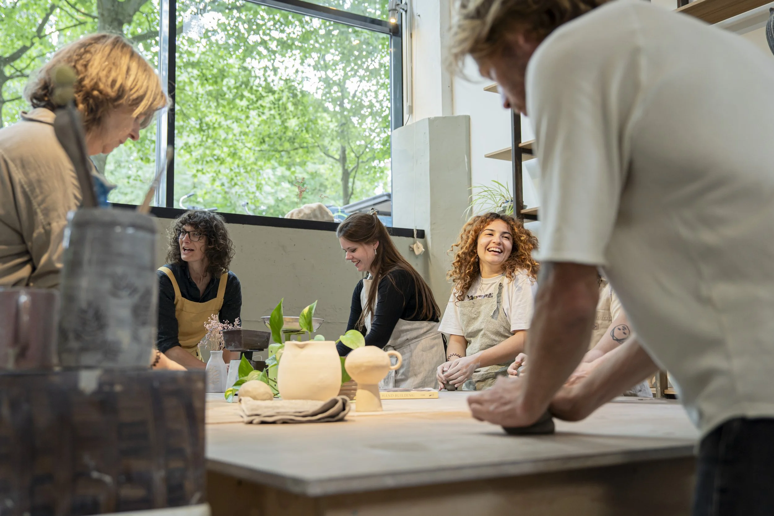 People participating in a pottery class, working on clay projects at a workshop table with pottery tools and plant decorations, inside a studio with large windows showing green trees outside.