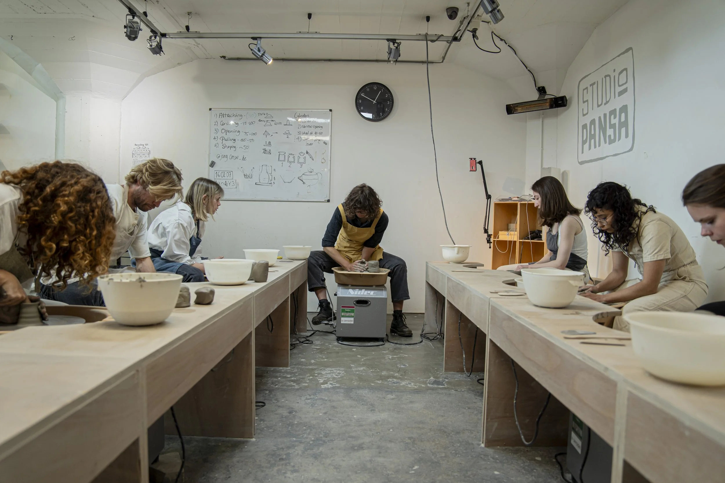 People working at pottery tables in a ceramic studio, with bowls, tools, and a whiteboard on the wall.