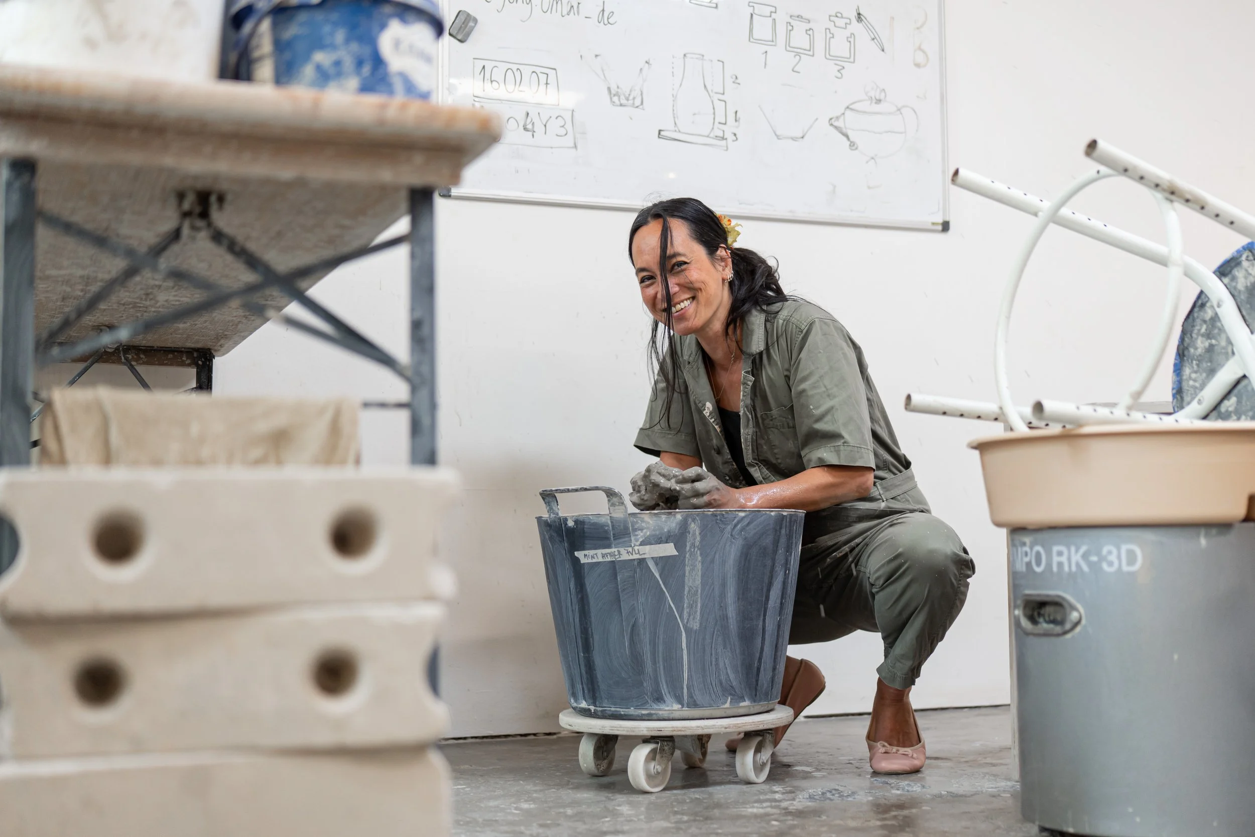 A woman crouched on the floor smiling, surrounded by buckets and pottery tools in a workshop. There is a whiteboard with sketches behind her.