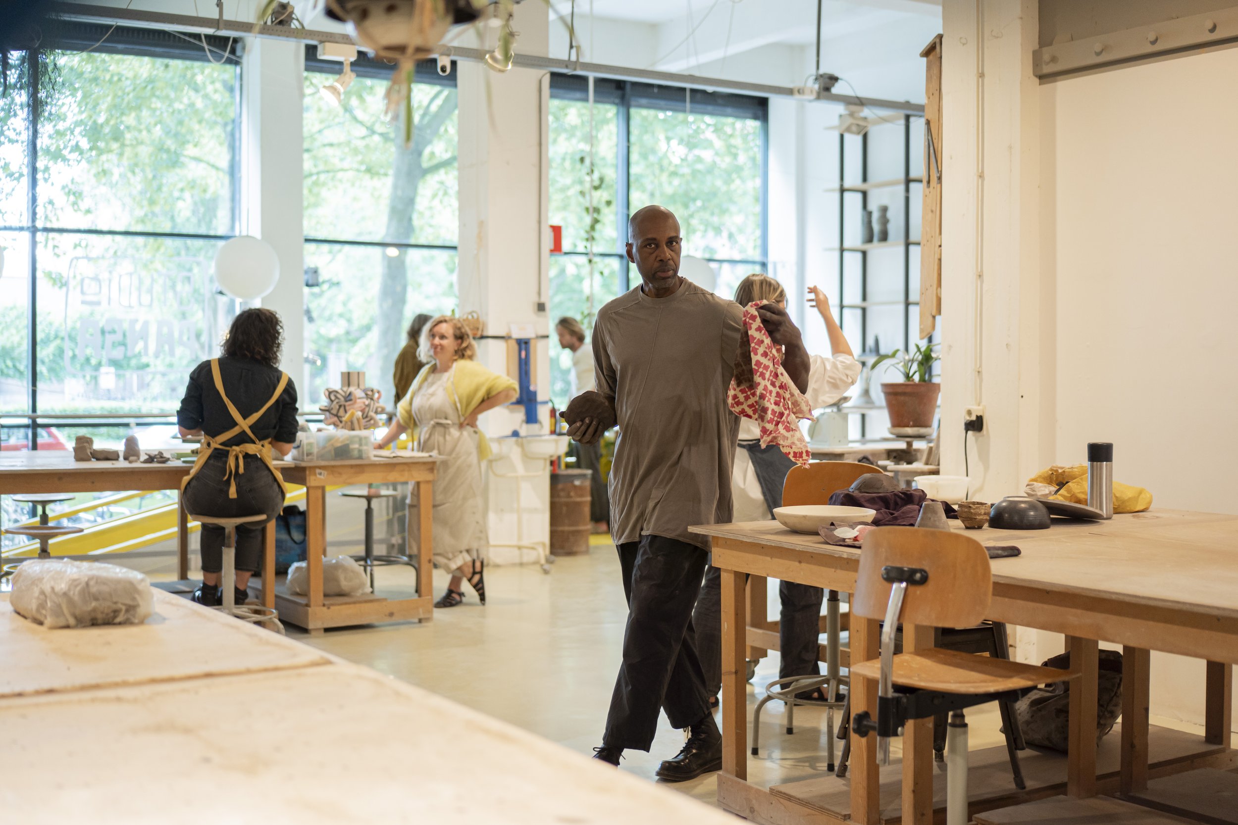 People working in a well-lit pottery studio with large windows, wooden tables, and shelves.