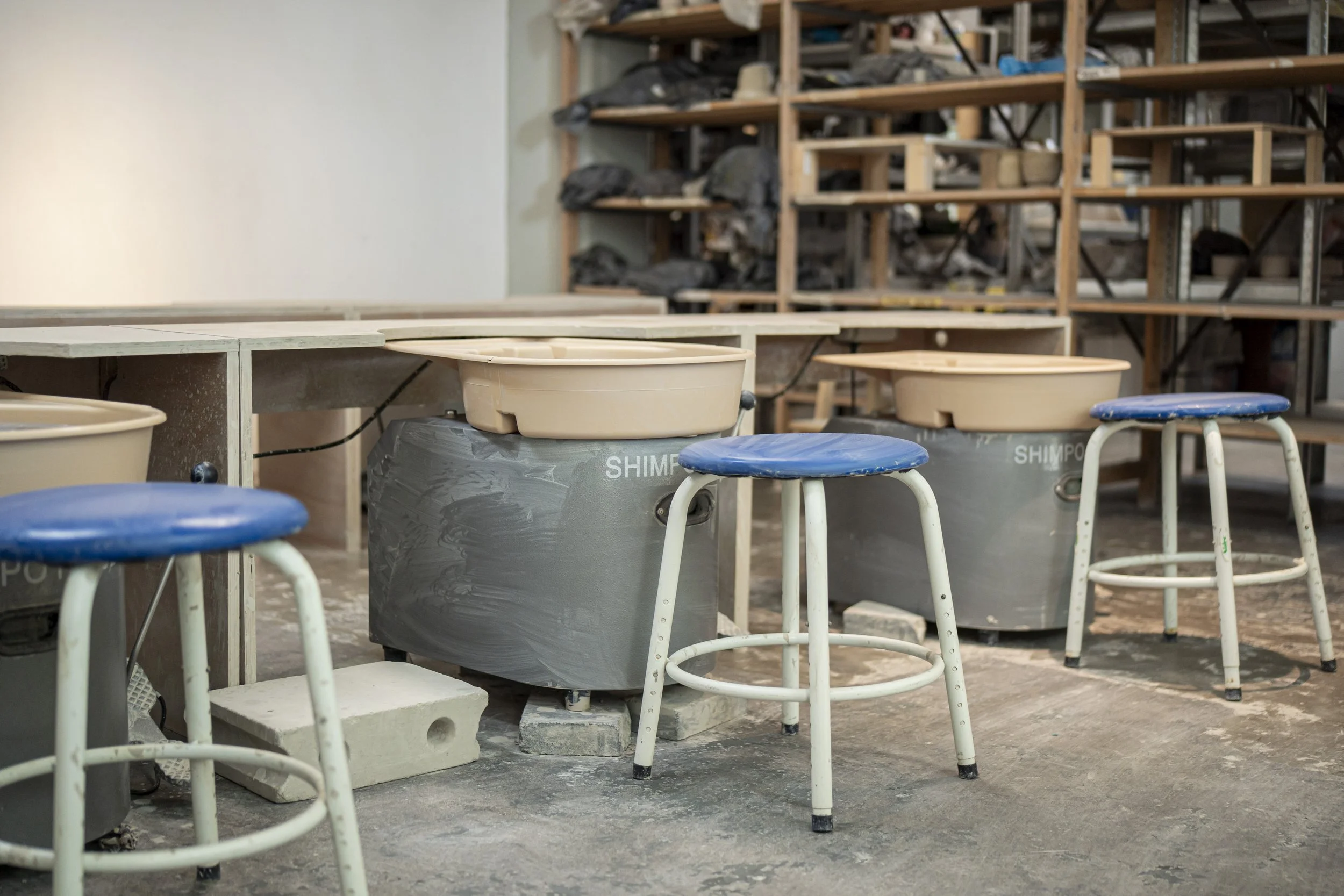 Ceramic pottery studio with three work tables, two blue and one beige, each covered with a beige pottery piece in progress. Three vintage, white stools with blue cushioned seats are positioned in front of the tables. In the background, there are wood