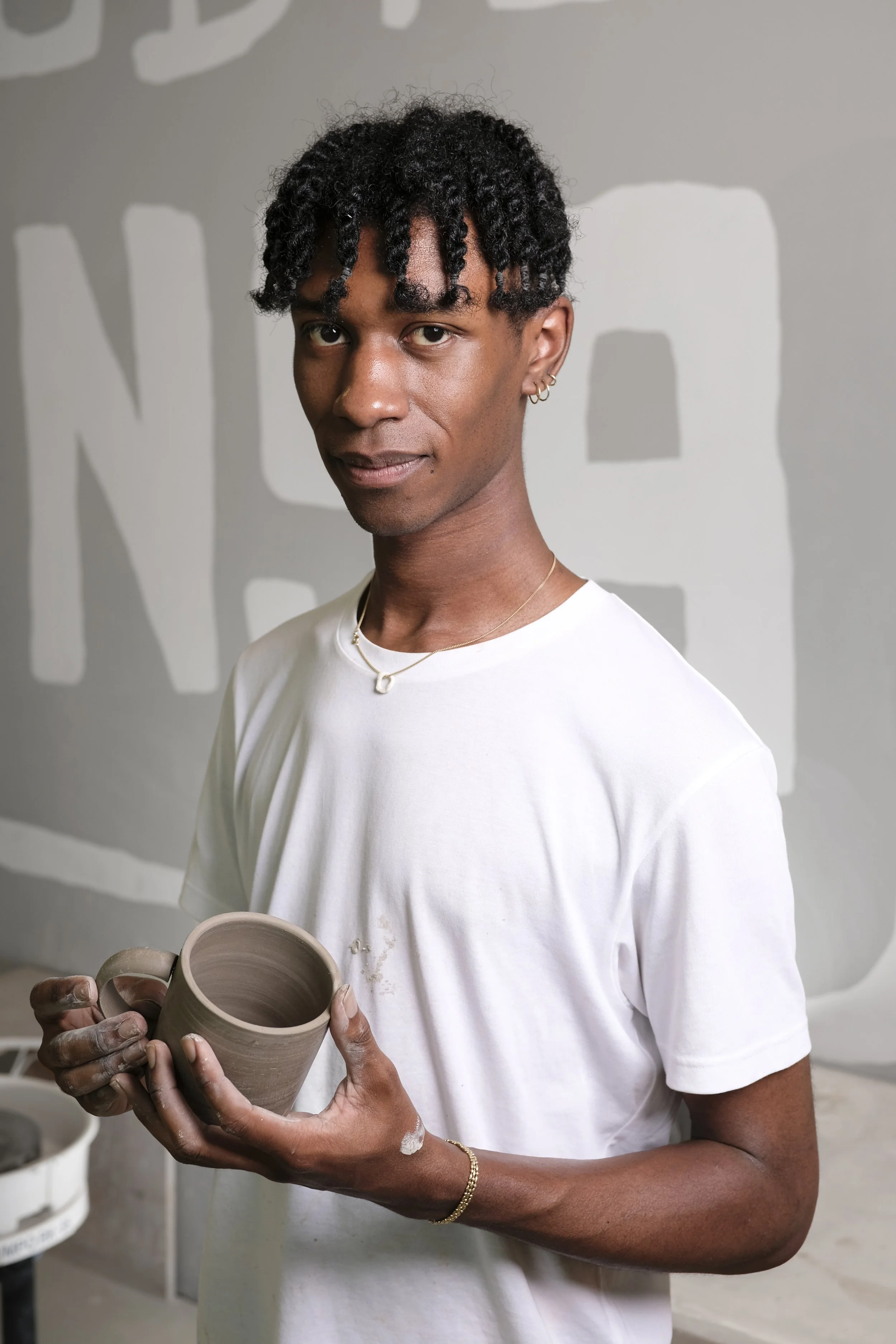 A young man with curly black hair and earrings, holding a ceramic mug, standing in front of a wall with abstract art. He is wearing a white t-shirt and a gold necklace and bracelet, with some clay residue on his hands and shirt.
