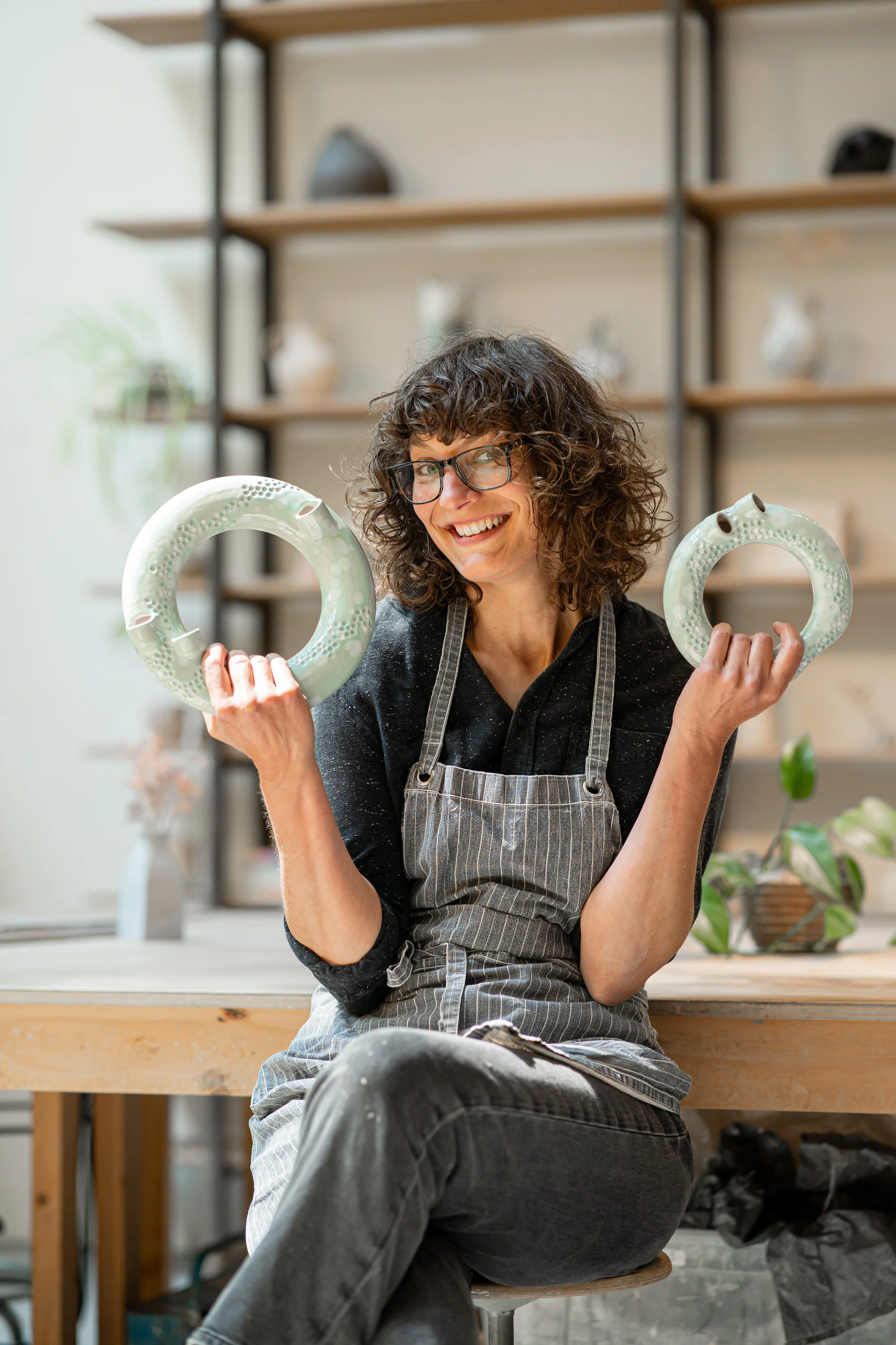 A woman with curly hair, glasses, wearing a black shirt and striped apron, smiling and holding two ceramic doughnuts in a pottery studio.
