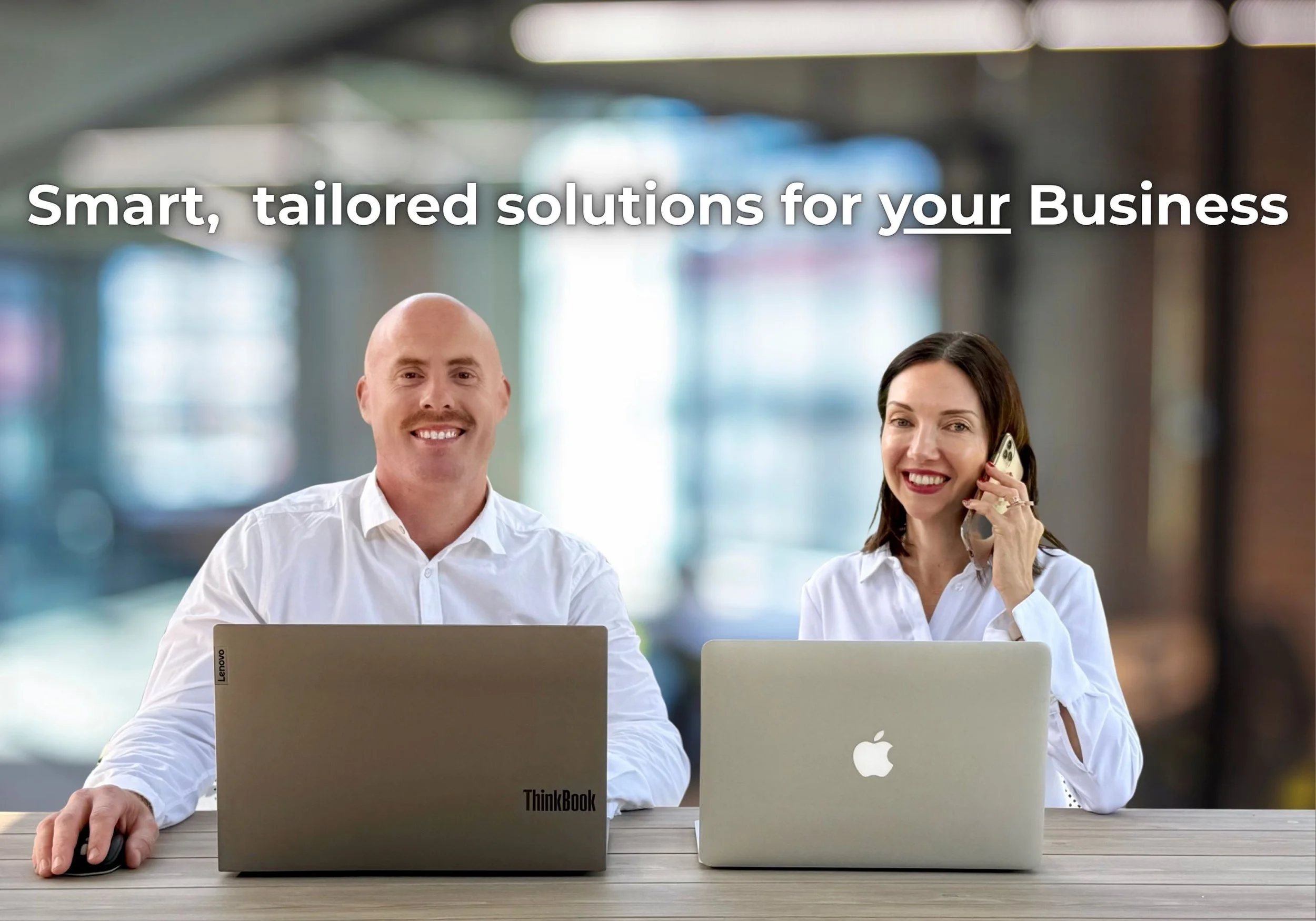 Two professionals, a man and a woman, sitting at a table with laptops in an office setting. The man has a shaved head and is smiling at the camera, using a Lenovo ThinkBook laptop. The woman has dark hair and is talking on her mobile phone, using a silver MacBook. The background is blurred with natural light coming through windows. Overlaid text reads, "Smart, tailored solutions for your Business."