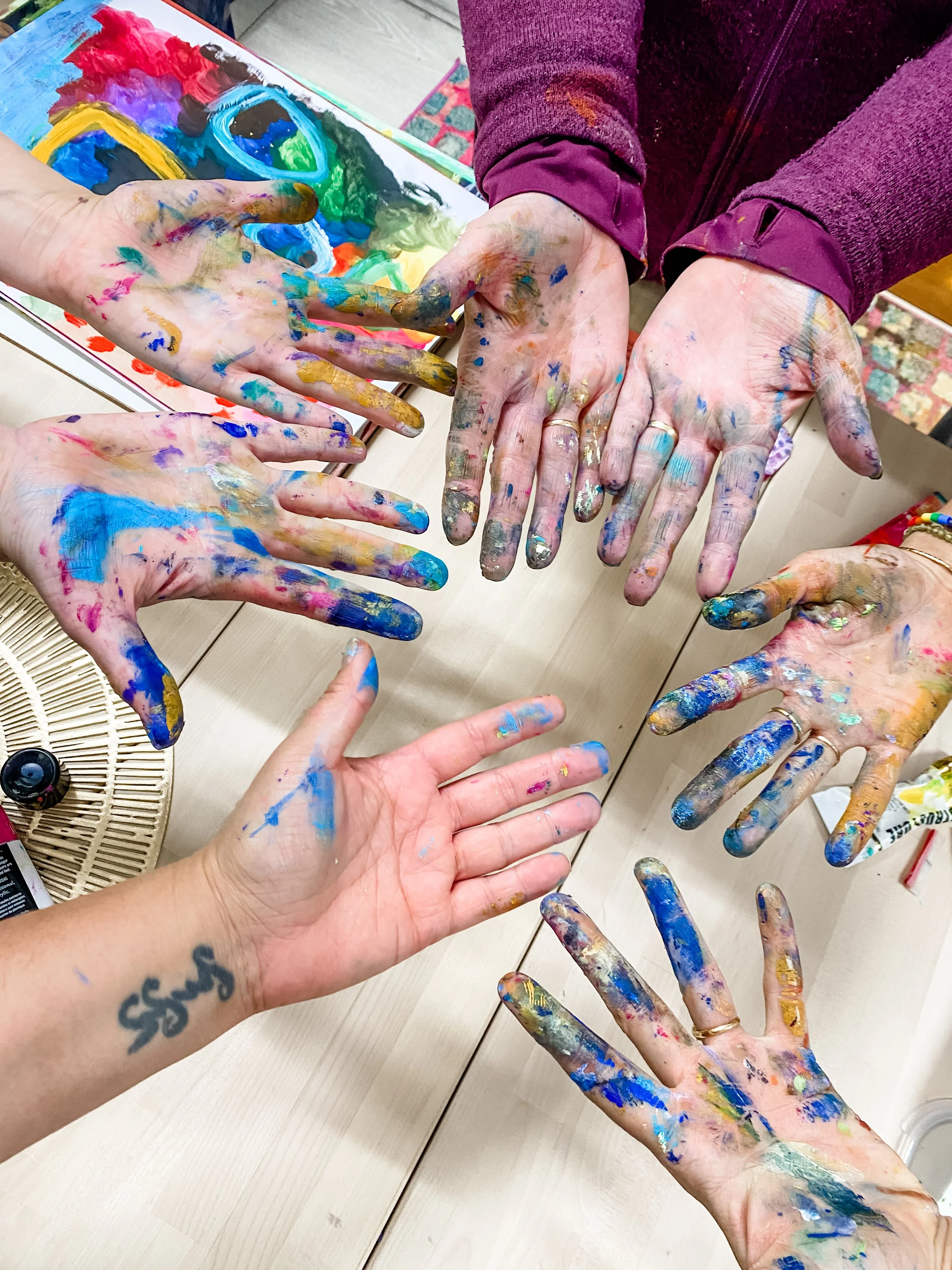 Multiple hands covered in colorful paint, with some hands showing rings and tattoos, and a colorful painted paper in the background.
