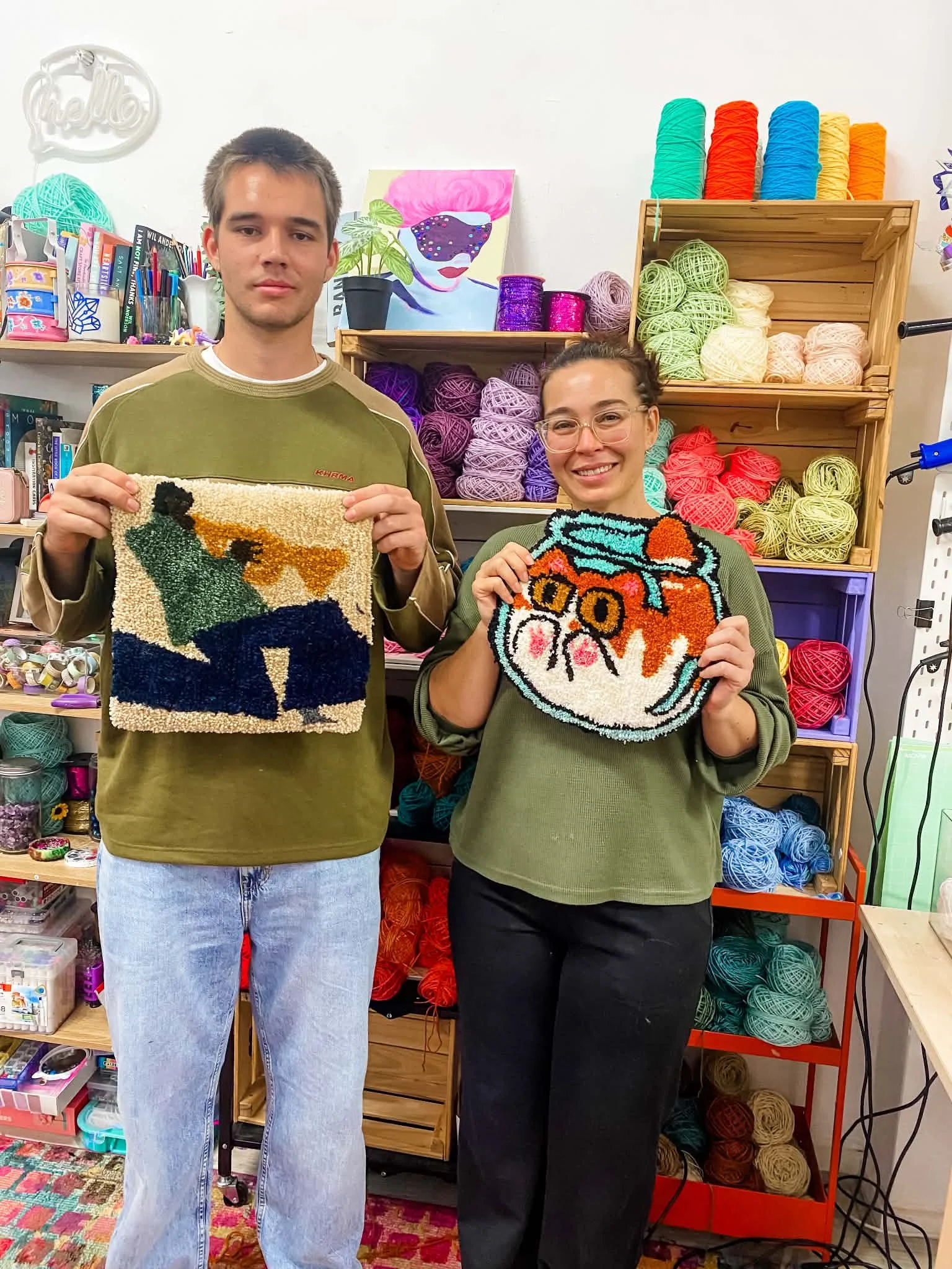 Two people standing in a craft store holding small colorful rugs, surrounded by yarn and craft supplies.