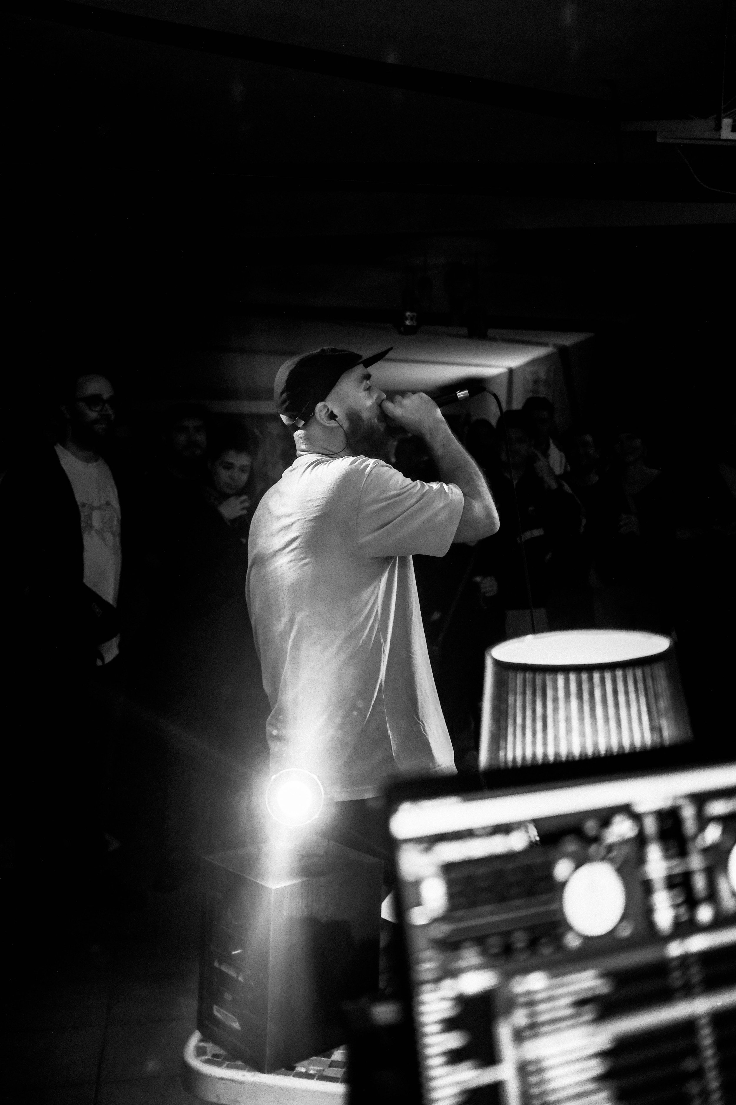 Ein Mann performt mit einem Mikrofon auf einer Bühne während einer Musikveranstaltung, umgeben von einem Publikum, im Schwarz-Weiß-Stil.