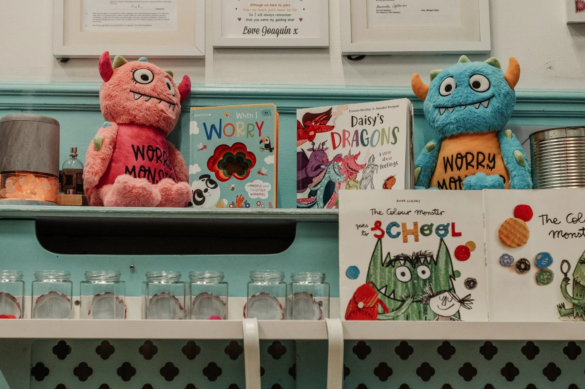 Two plush monsters, one pink with one eye and the other blue with two eyes, are placed on a bookshelf among children's books and artwork. The pink monster has the words 'WORRY MONSTER' on its front, and the blue monster has the same text.