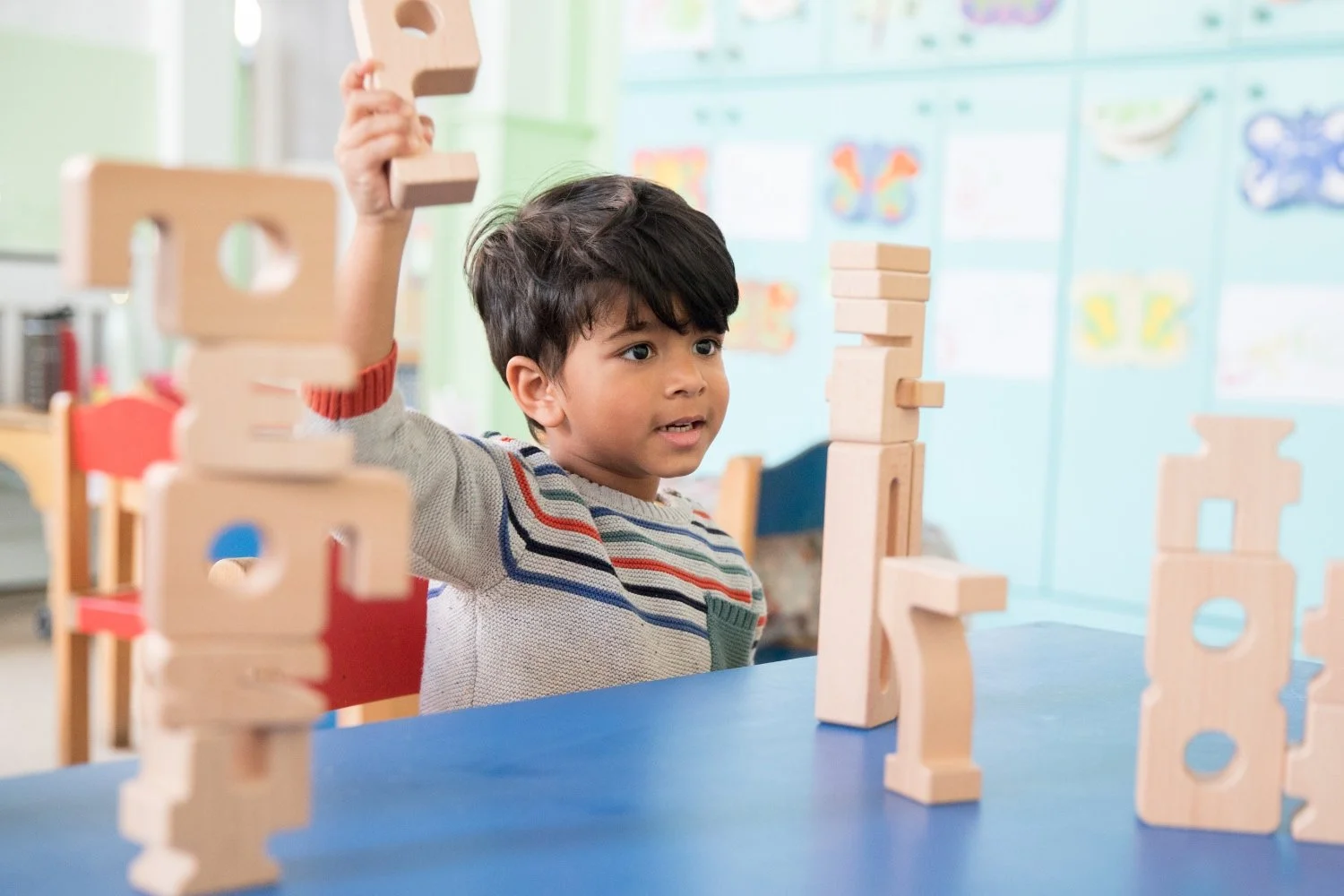 A young boy playing with wooden building blocks at a blue table in a classroom.