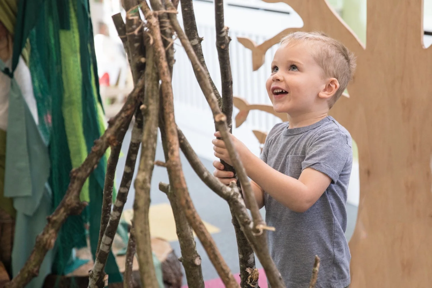 A young boy with short blond hair and a grey t-shirt is smiling and playing with wooden sticks arranged in a teepee or hut structure.