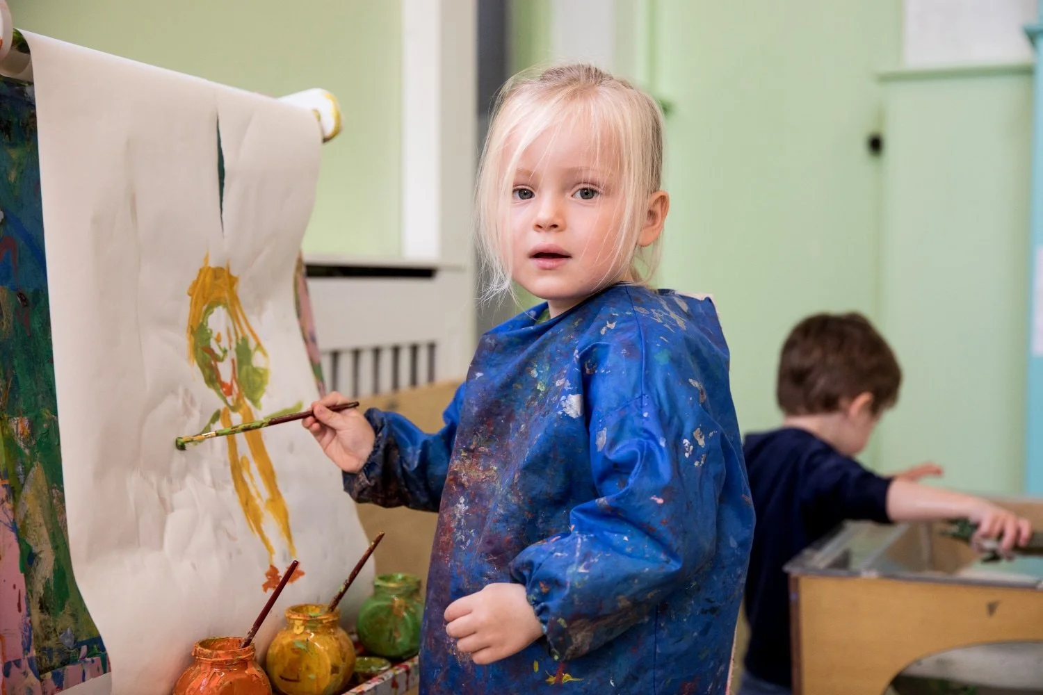 A young girl with blonde hair is painting on a large sheet of paper in an art classroom. She is wearing a blue painting smock stained with various colors of paint. A boy in the background is working at a table."