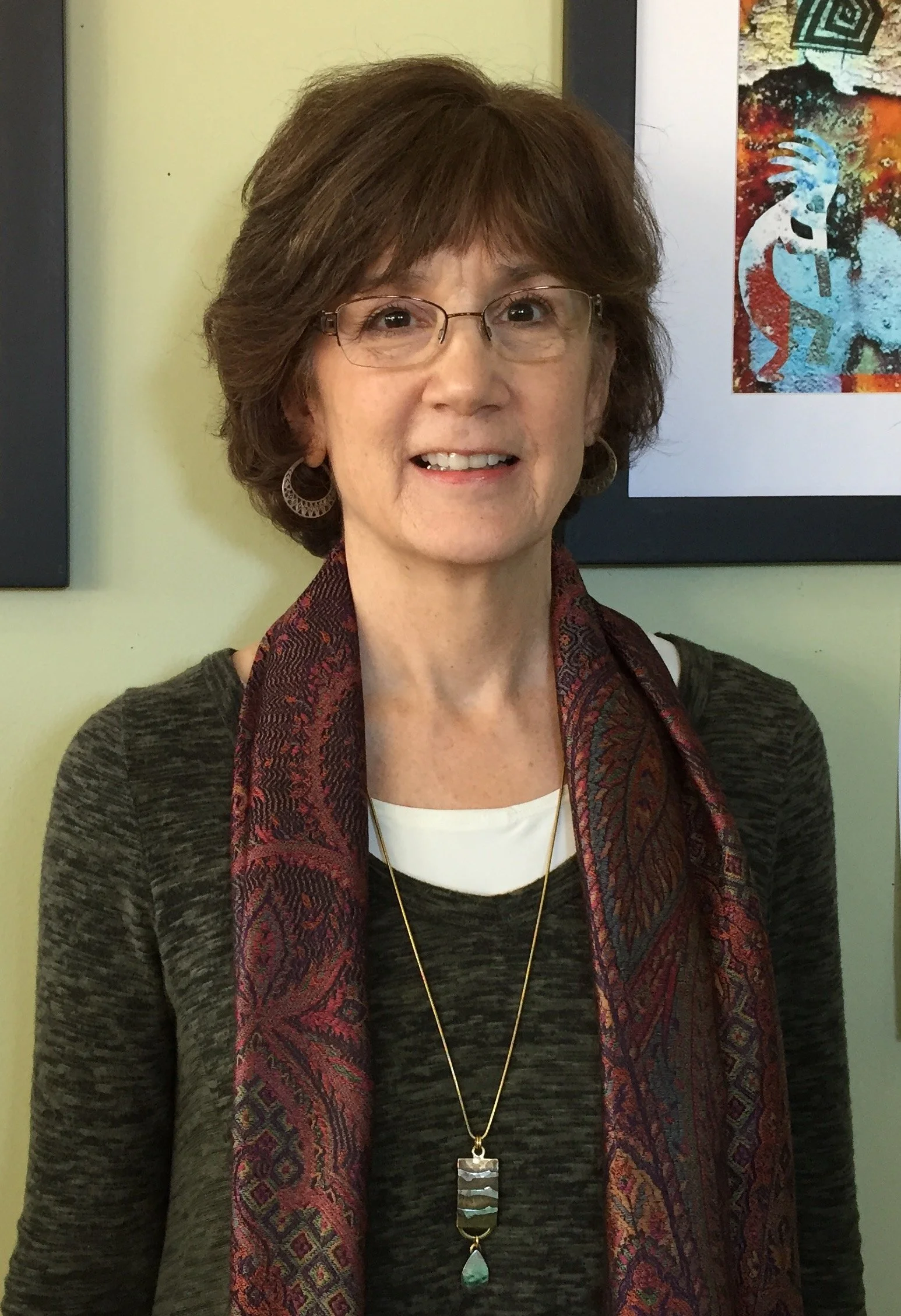 A smiling middle-aged woman with short brown hair, glasses, and earrings, wearing a patterned scarf and a dark sweater, standing in front of colorful artwork on a light-colored wall.