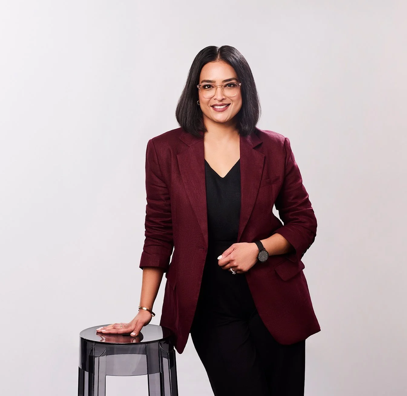 Woman with shoulder-length black hair, glasses, wearing a maroon blazer over black top, standing next to a small stool, smiling at the camera against a plain gray background.