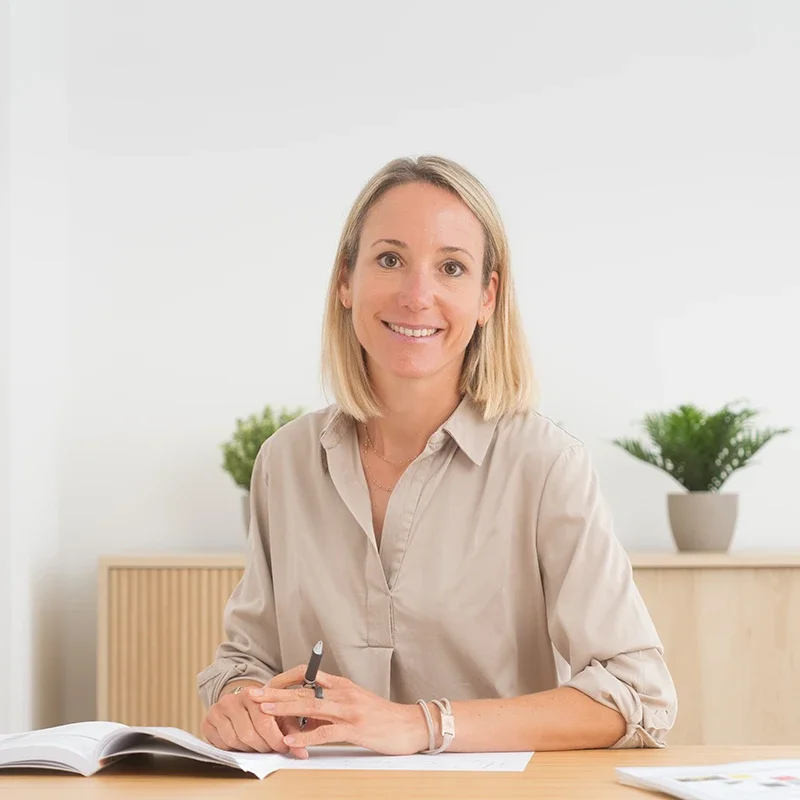 Eine lächelnde Frau mit blonden Haaren sitzt an einem Tisch in einem hellen Raum, mit offenen Büchern und Dokumenten vor ihr, im Hintergrund befinden sich zwei grüne Pflanzen in weißen Töpfen.