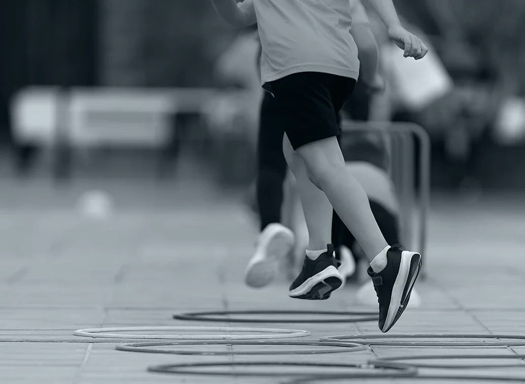 Zwei Schüler springen durch Hula-Hoop-Reifen auf einem Spielplatz, im Hintergrund sind Bänke und Fahrzeuge sichtbar.