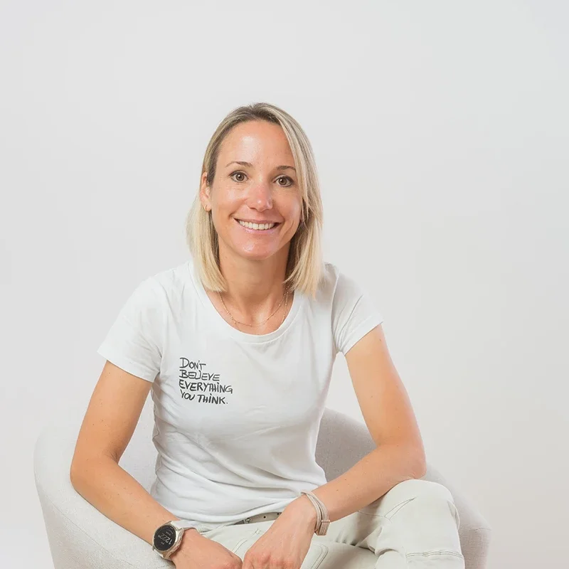 Eine lächelnde blonde Frau sitzt auf einem weißen Stuhl vor einer weißen Wand, trägt ein weißes T-Shirt und eine schwarze Uhr.
