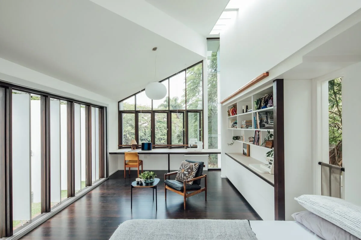 An airy modern room with dark wood floors, expansive windows framing lush outdoor greenery, an integrated workspace, a cozy armchair, and white built-in shelving.