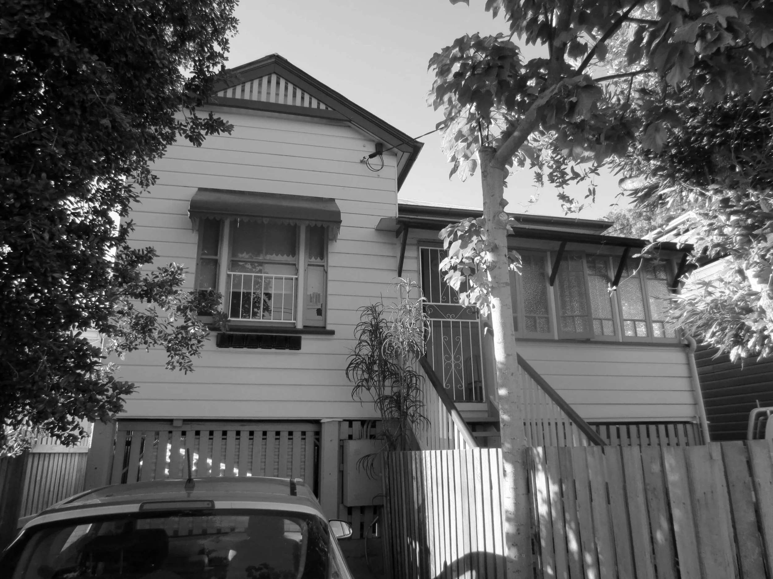 A monochrome elevated Queenslander house, with a gabled roof and an ornate awning, is partially visible through dense trees, with a car's roof in the foreground and a wooden fence on the right.