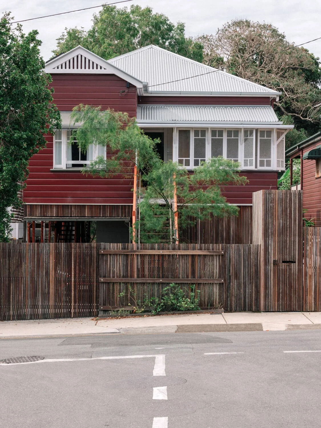 A maroon elevated house with a corrugated metal roof and an enclosed front veranda, featuring white windows, stands behind a dark wooden fence on a residential street.