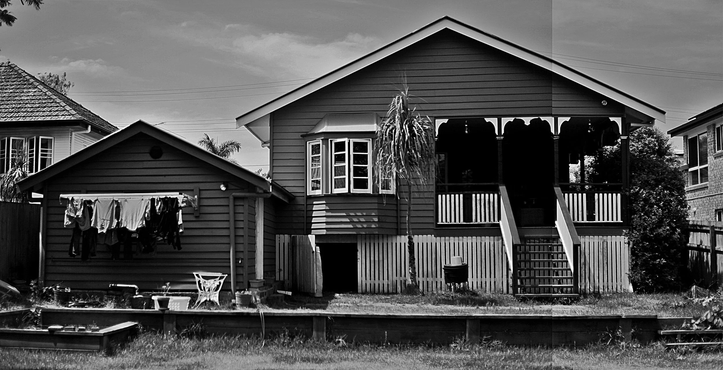 In black and white, a traditional high-set wooden house with a verandah stands next to a smaller shed where laundry hangs to dry, surrounded by a grassy garden.