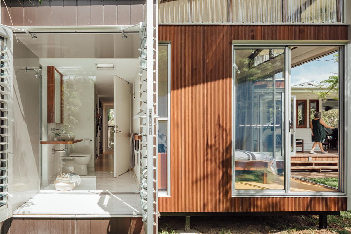 A modern home features an open white bathroom with louvered walls on the left, contrasting with a wood-paneled exterior and large glass doors revealing a bedroom and a person walking on an outdoor deck.