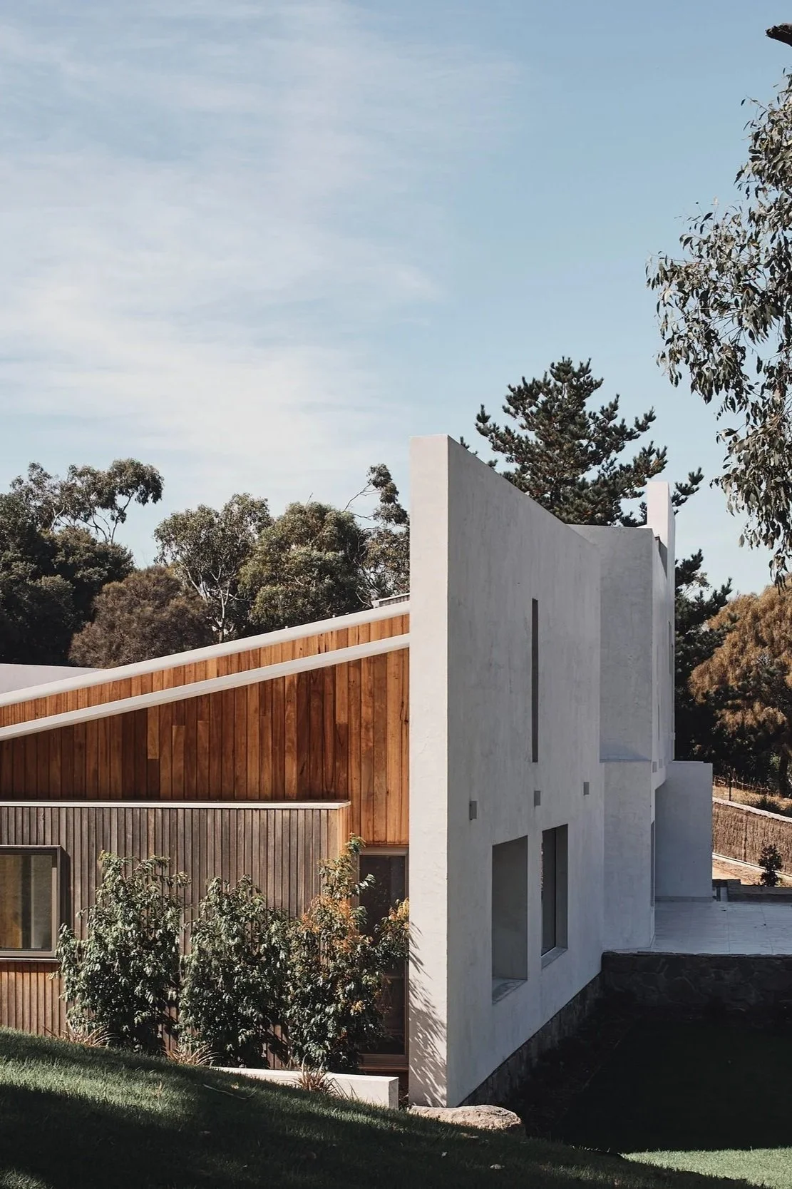 Modern house with a combination of wooden and white concrete exterior walls, surrounded by trees and a grassy area under a blue sky.