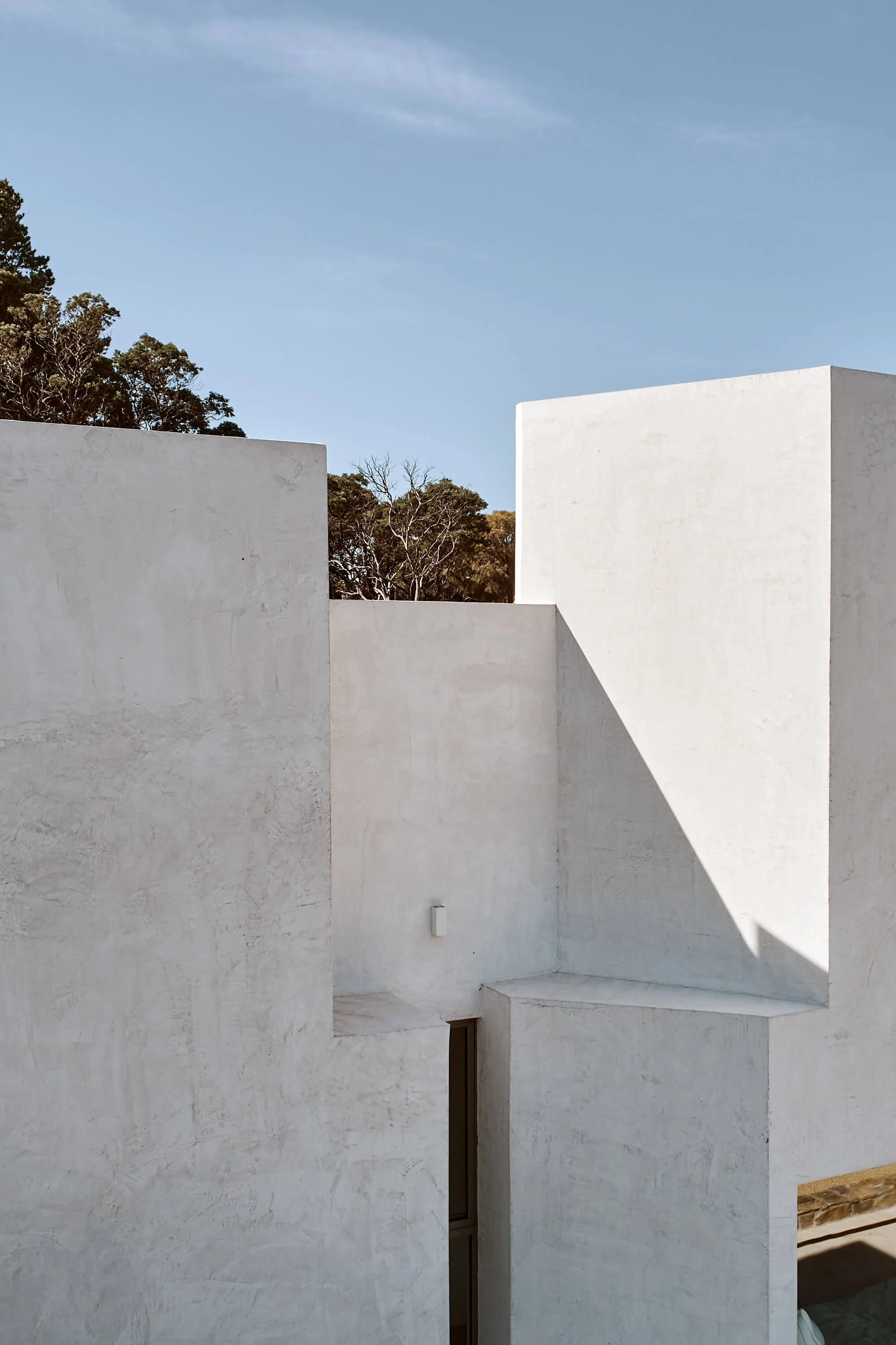 Geometric, textured concrete walls cast sharp shadows under a clear blue sky, with lush green trees peeking over and through the minimalist structure.
