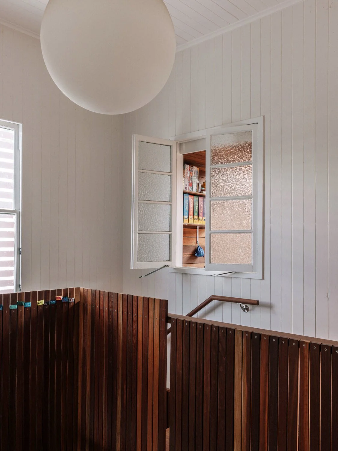 A bright interior landing features white paneled walls, a large spherical light fixture, and a dark wooden slatted balustrade, with an open frosted window revealing a bookshelf in an adjacent room.