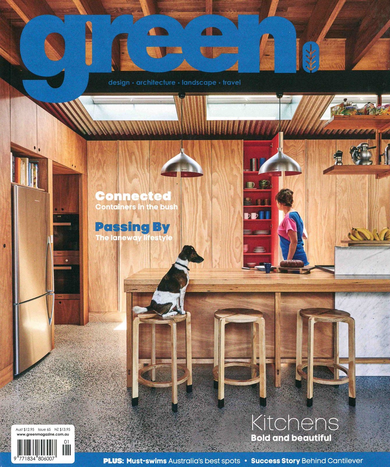 Interior of a modern kitchen with wooden cabinets, a marble countertop, and bar stools. A dog sits on a stool at the island, and a woman is standing nearby. The magazine cover features the title 'green' and highlights topics like design, architecture, landscape, and travel.