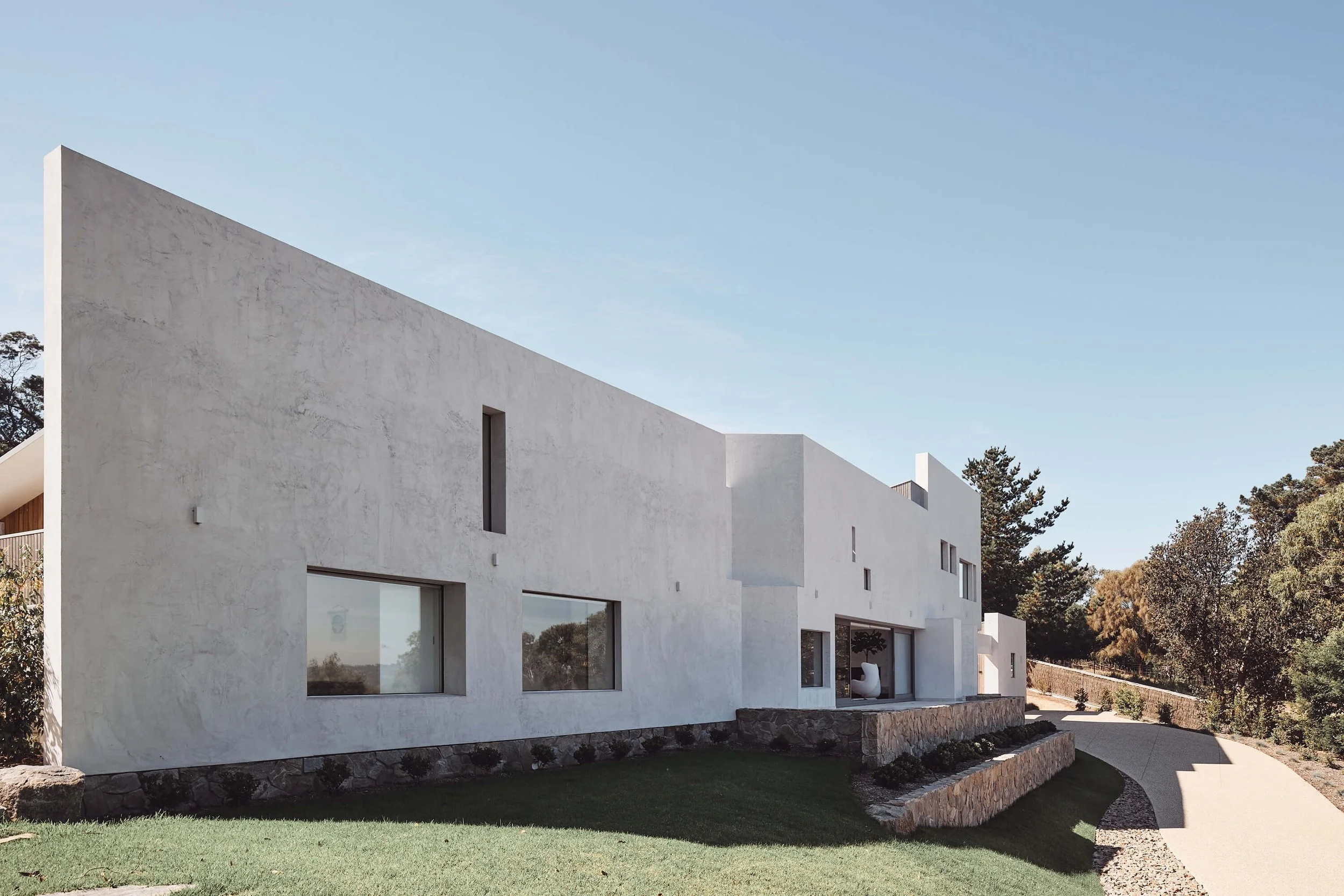 A striking modern house with a textured white stucco facade and geometric angles stands on a terraced green lawn, flanked by a winding driveway and dense trees under a clear sky.