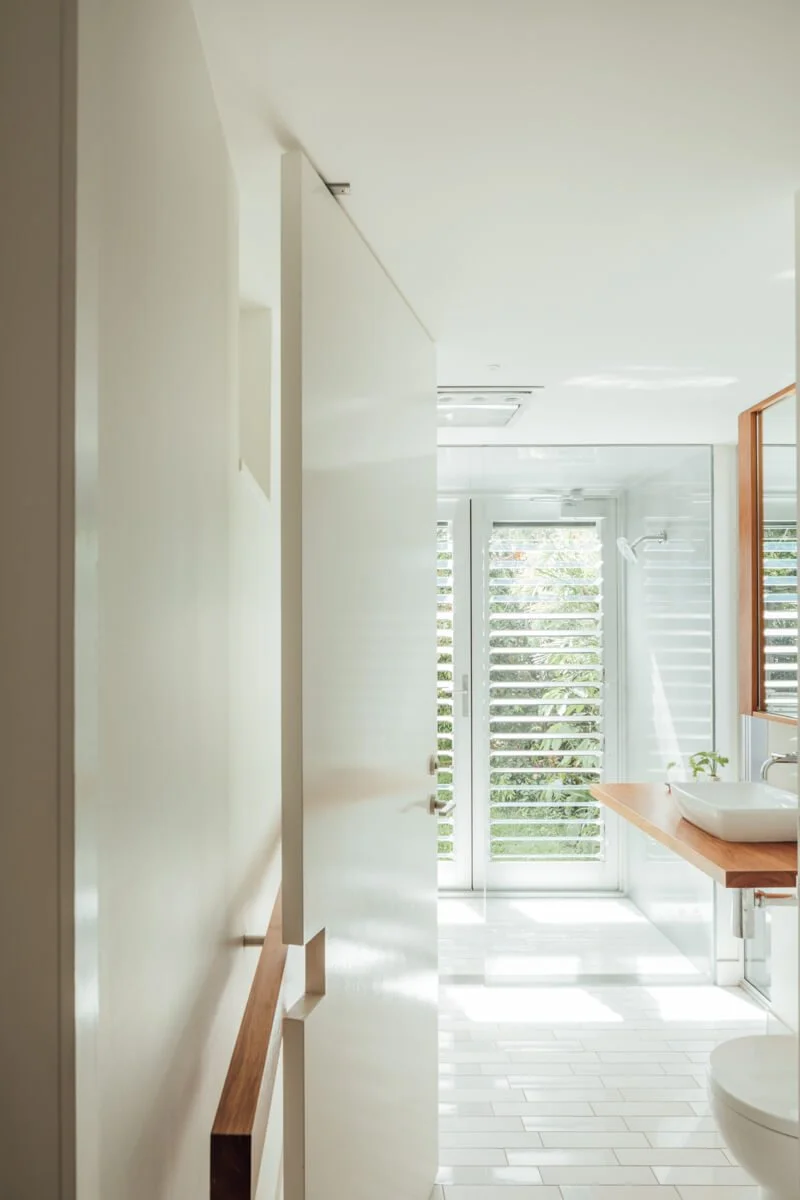 A minimalist modern bathroom features white subway tile floors, a long floating wooden vanity with a white vessel sink, a glass shower, and a large louvered window revealing green foliage outside.