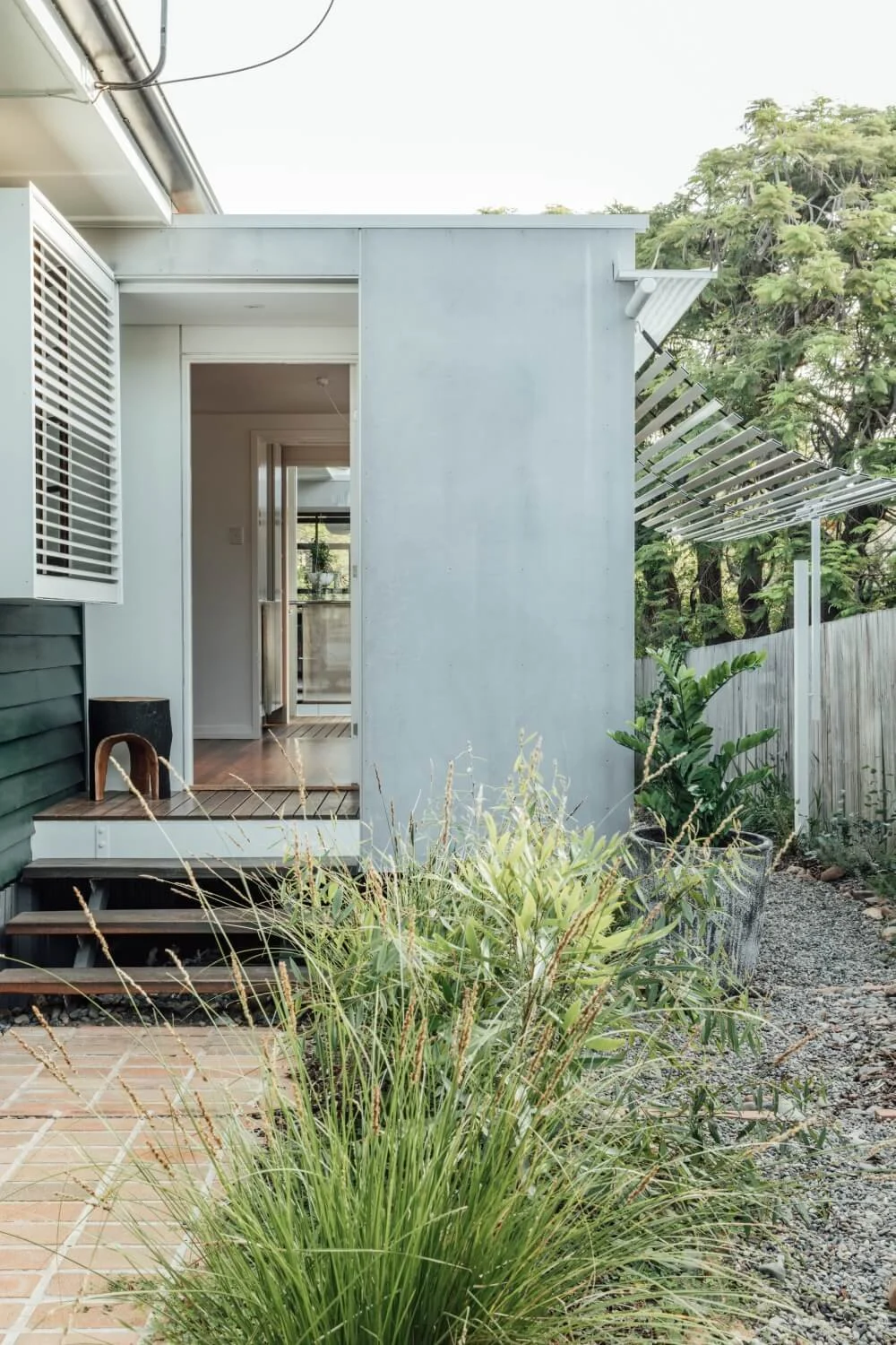 A modern house entrance with wooden steps and a deck, framed by lush green landscaping and a gravel path leading to a white slatted pergola.