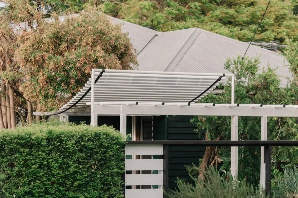 A dark-sided house with a grey roof is partially visible behind a white pergola topped with a retractable striped awning, framed by a dense green hedge and various trees.