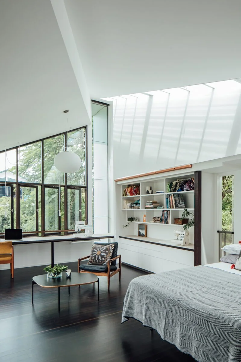 A bright, modern open-plan room with high ceilings, large windows overlooking lush trees, furnished with a workspace, built-in bookshelves, an armchair, and a bed with a patterned blanket.