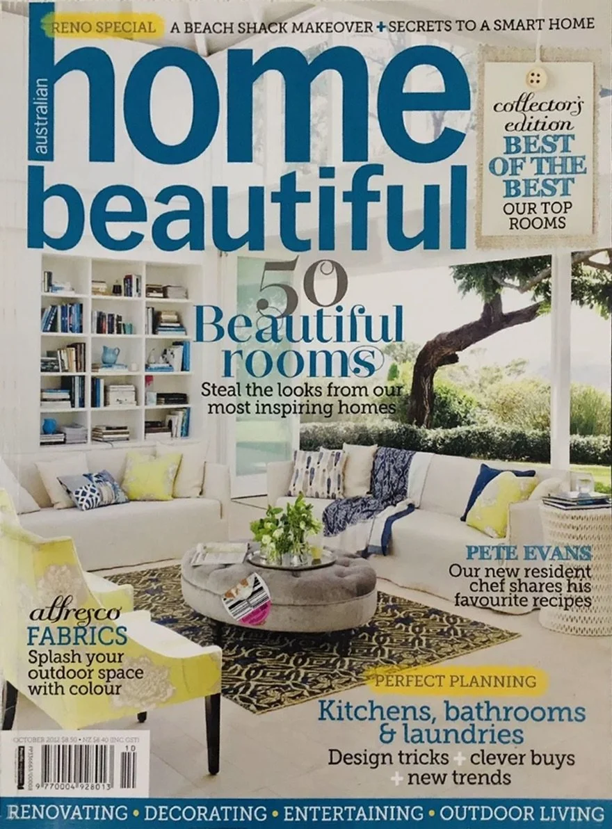 Front cover of 'Home Beautiful' magazine featuring a living room with white sofas, yellow and blue pillows, a round ottoman with a plant on top, a bookshelf, large window with a view of greenery and a tree outside.