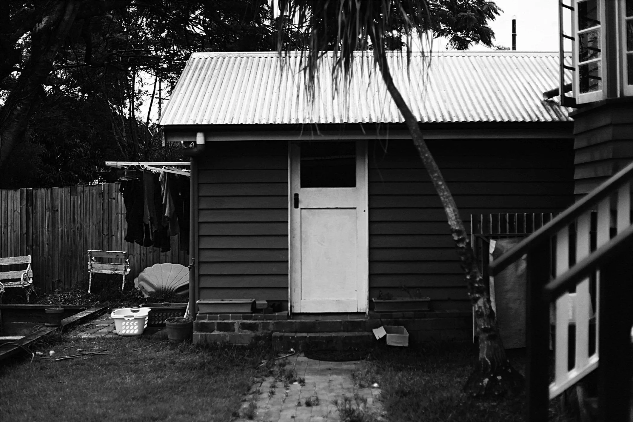 A weathered shed with a white door and corrugated iron roof stands in a monochrome backyard, surrounded by drying laundry, a wooden fence, various garden items including a bench, and dense trees, with a house and railing visible on the right.