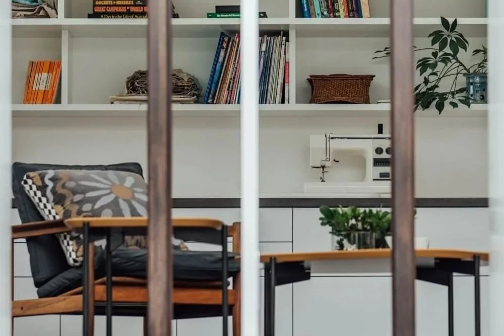 A modern interior featuring white bookshelves filled with books and plants, a sewing machine on a counter, and an armchair with a patterned cushion, all viewed through vertical room dividers.