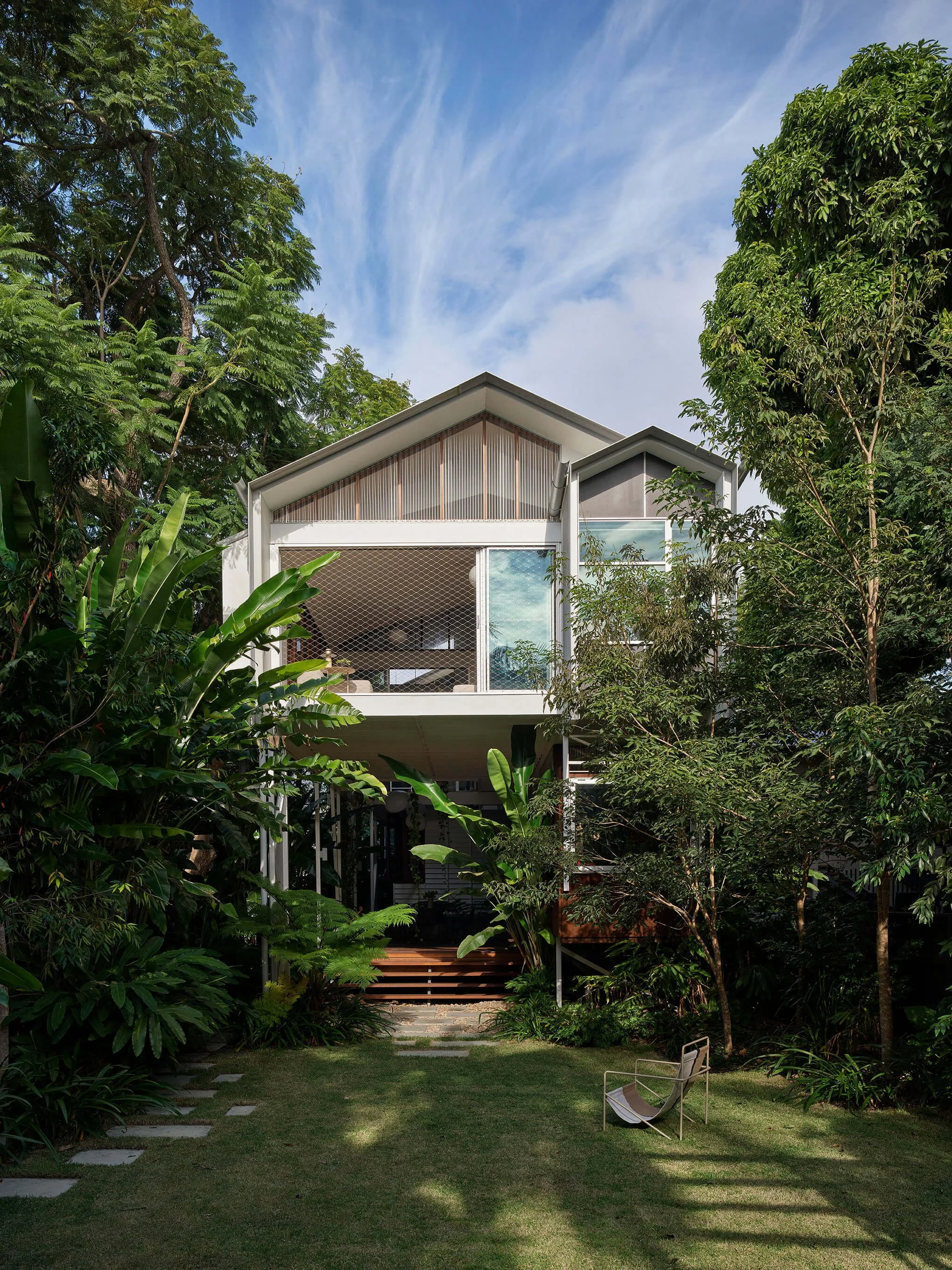 Modern two-story house surrounded by lush greenery and trees, with a grassy yard and a single chair in the foreground, under a blue sky with wispy clouds.
