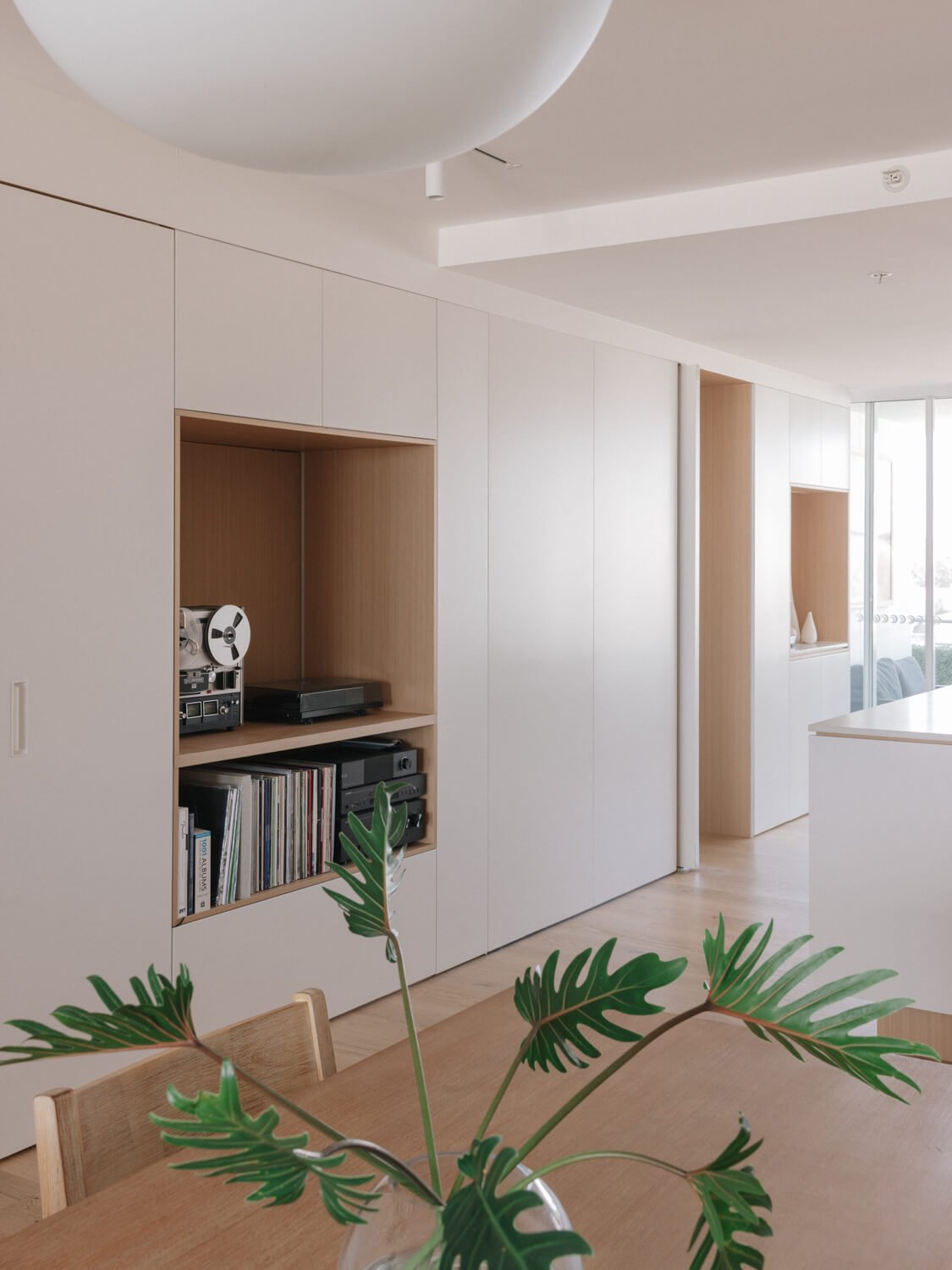 A modern, bright interior living space with white walls and a light wood coffee table. A plant with green, split leaves is on the table. There are built-in shelves in the wall holding vinyl records, books, and stereo equipment. Large windows let in natural light.