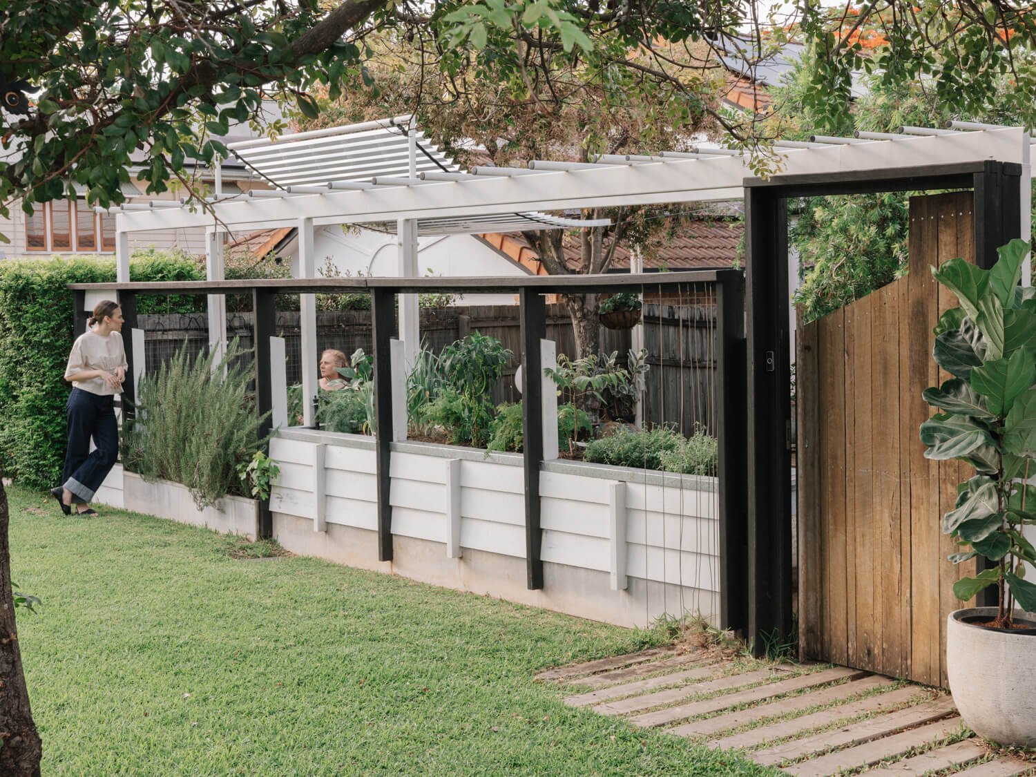 Two women engage with lush plants in white raised garden beds, beneath a dark-framed pergola and beside a rustic wooden gate in a sunny backyard.