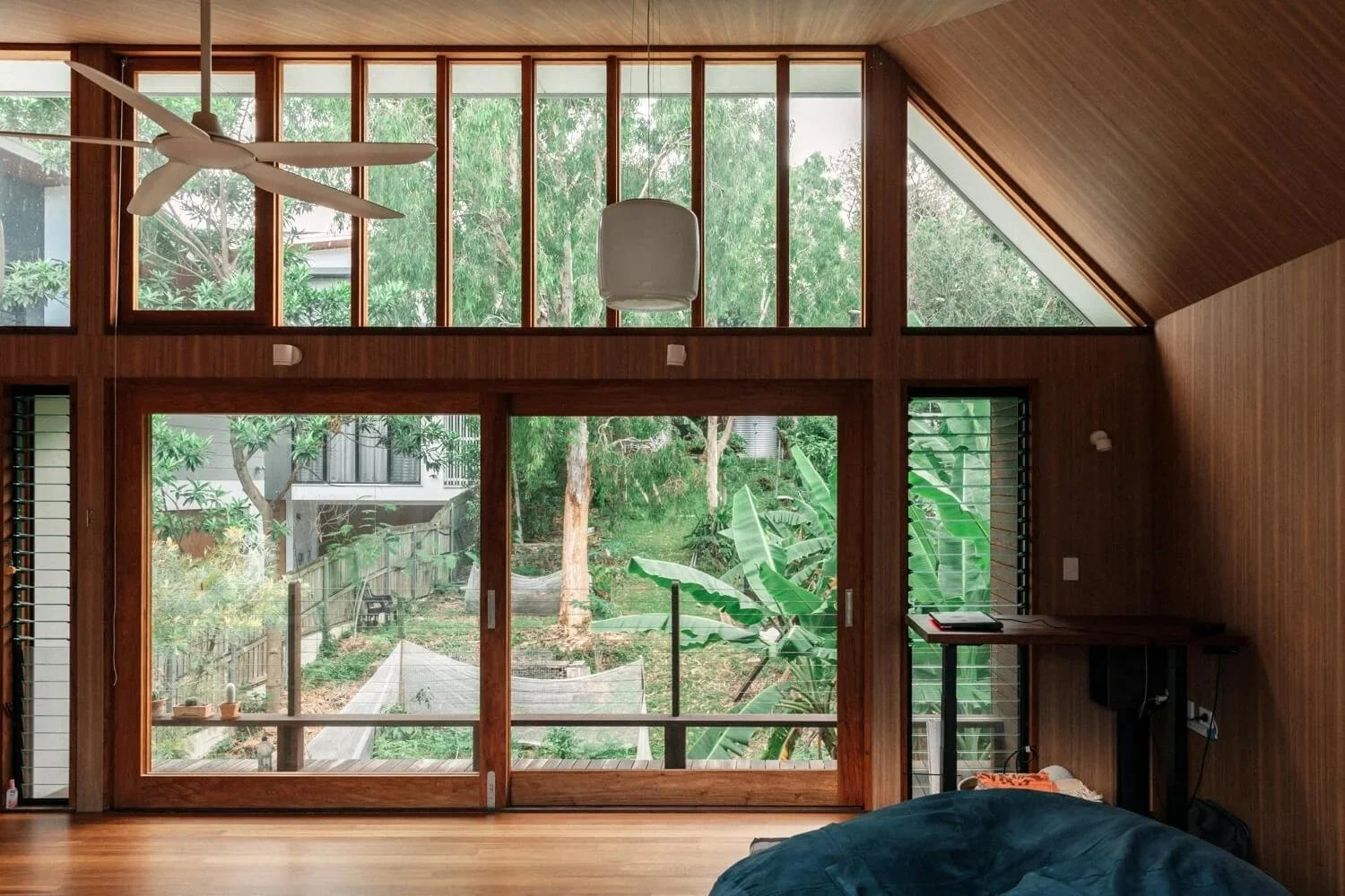 A modern room features warm wood paneling, a vaulted wood ceiling with a white fan, and expansive glass doors and windows overlooking a lush, tropical garden.