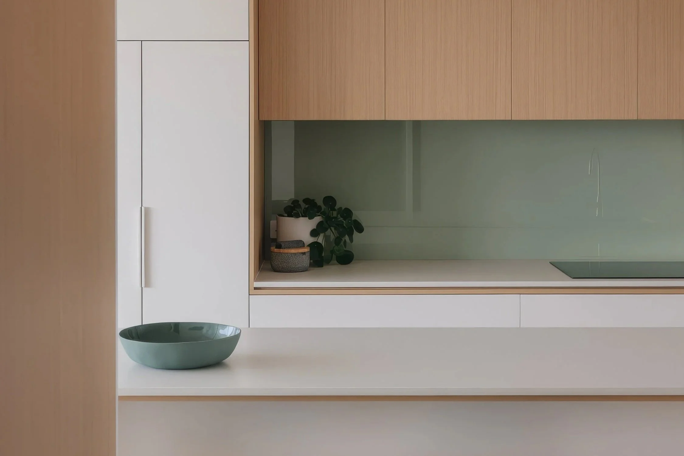 A clean, modern kitchen features light wood upper cabinets, white lower cabinets and counter, a pale green backsplash, and a teal bowl on the counter.