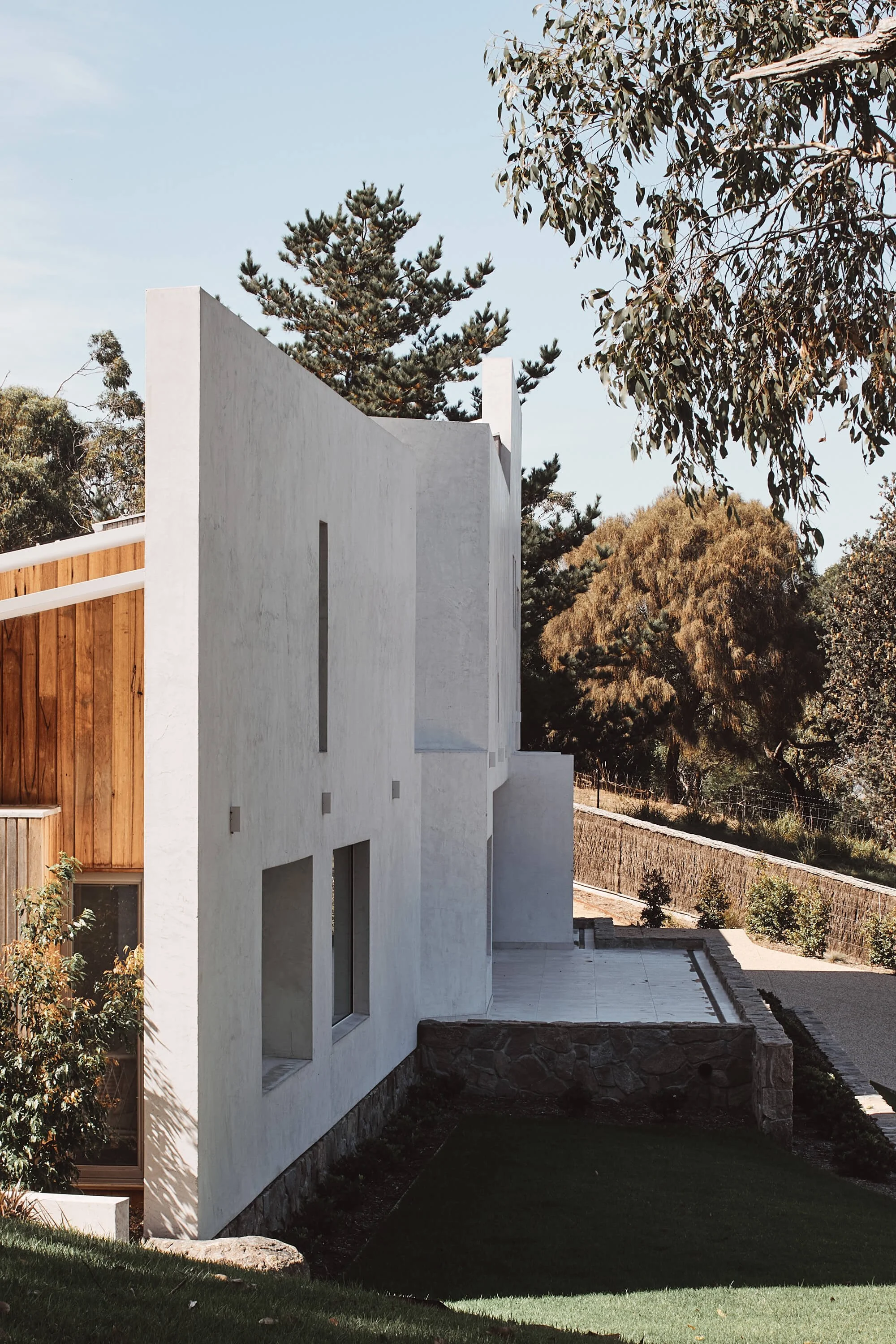 A striking modern house with stark white walls and warm wooden panels nestles into a green, tree-filled hillside under a bright sky.