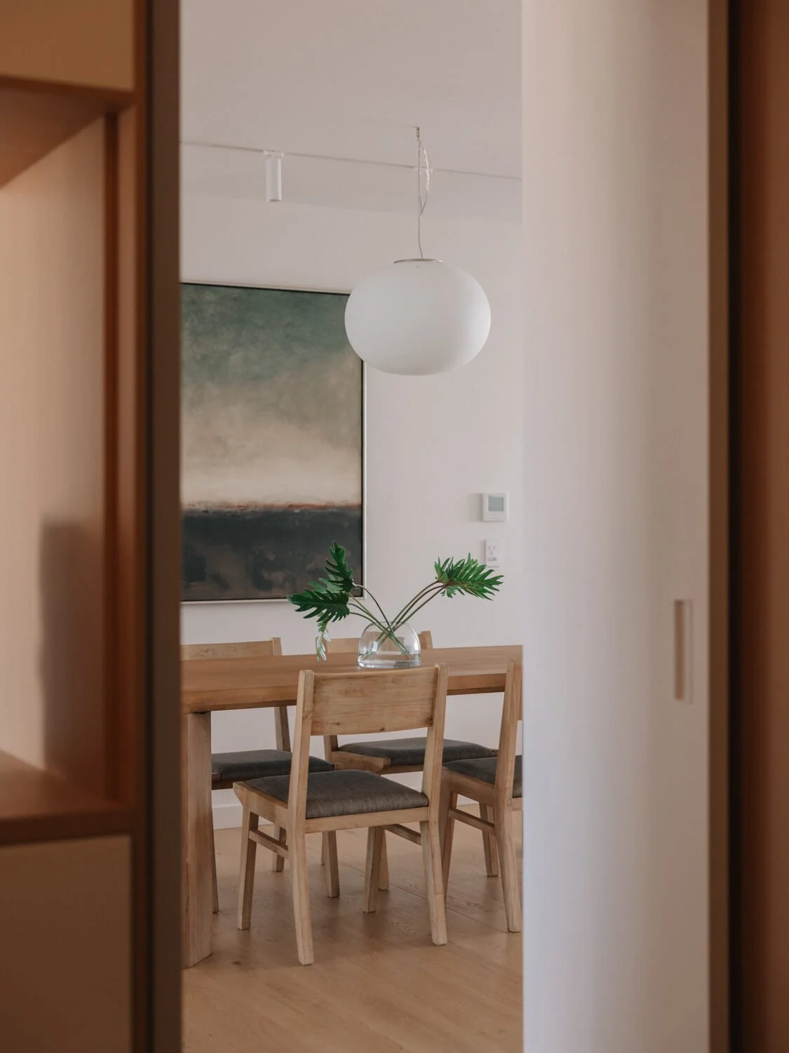 A modern dining room seen through an interior doorway features a wood table, chairs, a spherical pendant light, an abstract painting, and a vase of greenery.