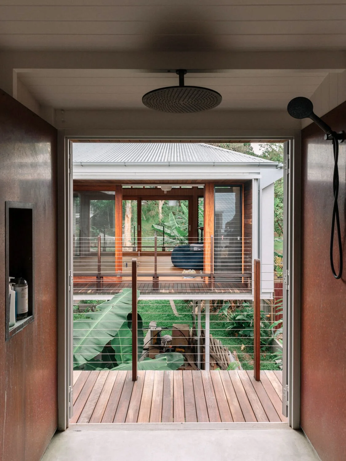 A modern outdoor shower with a visible rain head looks out onto a lush tropical garden featuring multi-level wooden decks and a contemporary house.