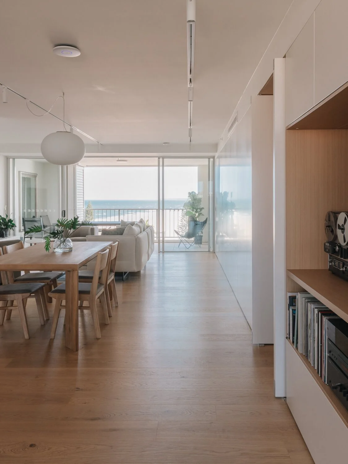 A bright, modern open-plan apartment with light wood floors features a dining area, living room, built-in shelving, and a large balcony overlooking the ocean.