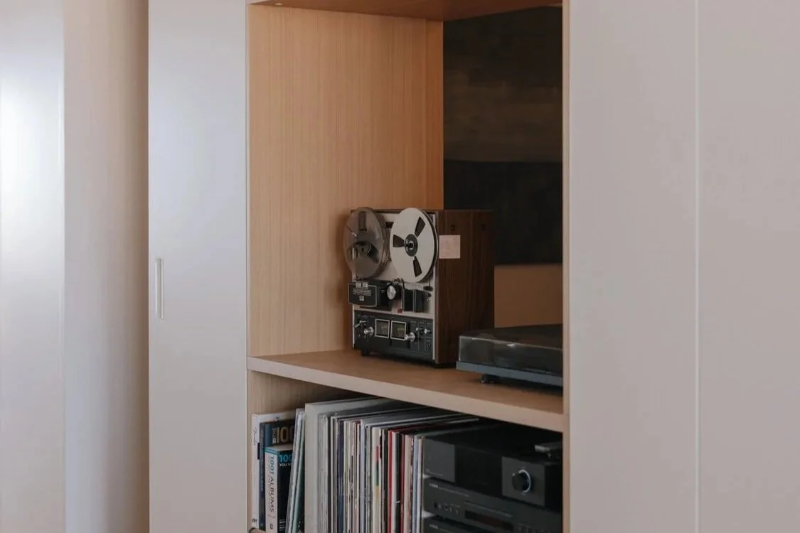 A vintage reel-to-reel tape recorder and modern audio equipment are displayed on light wood shelves, with vinyl records and books below, within a built-in cabinet.